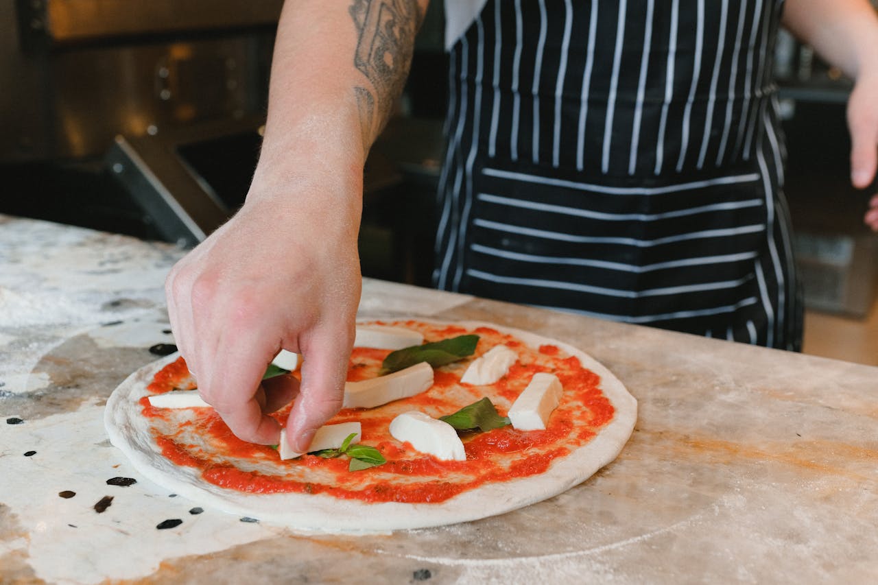 Close-up of a chef's hand adding ingredients to an uncooked pizza on a marble countertop.